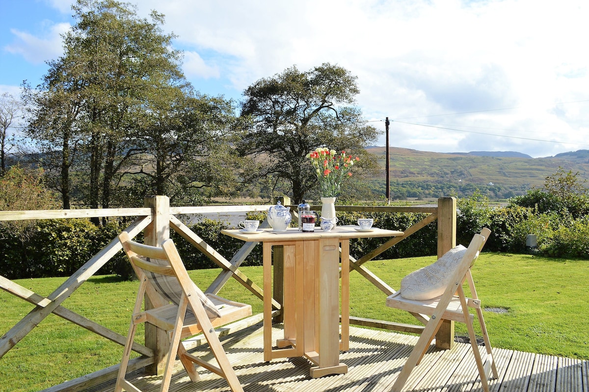 An outdoor deck area is presented with a wooden table set for two, featuring a vase of flowers. Two chairs are positioned beside the table, with views of the surrounding greenery and hills in the background. The sky is partly cloudy, adding to the serene atmosphere.
