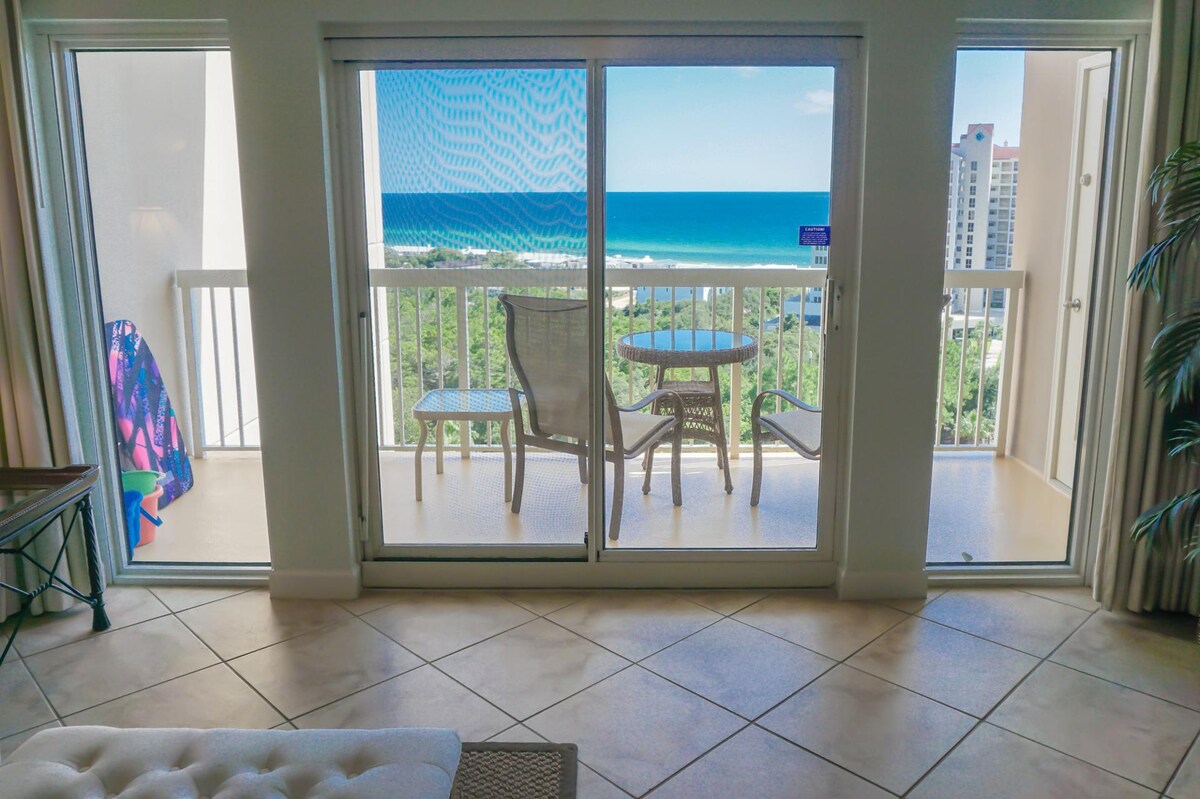 A view of the balcony with a table and chairs overlooking the Gulf of Mexico. Light blue water and clear skies are visible, framed by sliding glass doors. A colorful beach towel hangs on the railing, complementing the coastal atmosphere.