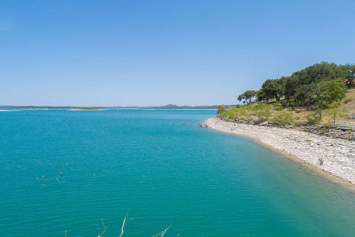 A scenic view of a tranquil lake with clear turquoise water, surrounded by rocky shorelines and lush greenery. The shoreline is dotted with trees, offering a peaceful landscape under a clear blue sky.