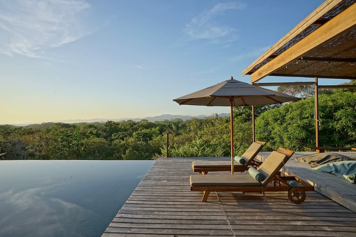 A serene infinity pool is featured alongside wooden loungers beneath a shaded umbrella, offering expansive views of the lush jungle and distant mountains. The calming water reflects the clear blue sky, blending seamlessly with the surrounding nature.