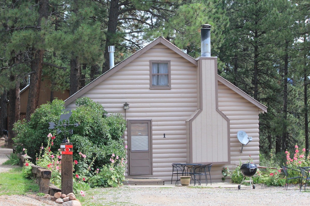 The exterior of the cabin is shown, featuring a beige log design with a prominent chimney. A covered entry door is visible, accompanied by a small table and chairs located in front. Lush green shrubs and flowers flank the sides, and tall trees provide a natural backdrop.