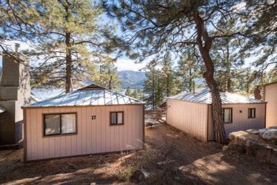 Two charming cabins are nestled among tall trees, showcasing light-colored exteriors with metal roofs. The surrounding natural landscape features a backdrop of mountains and a glimpse of the lake, enhancing the tranquil setting.
