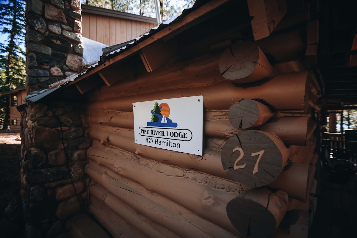 The exterior of the partially log cabin features a wooden sign reading "Pine River Lodge #27 Hamilton." The visible logs are complemented by a stone section, with a natural setting surrounded by trees.
