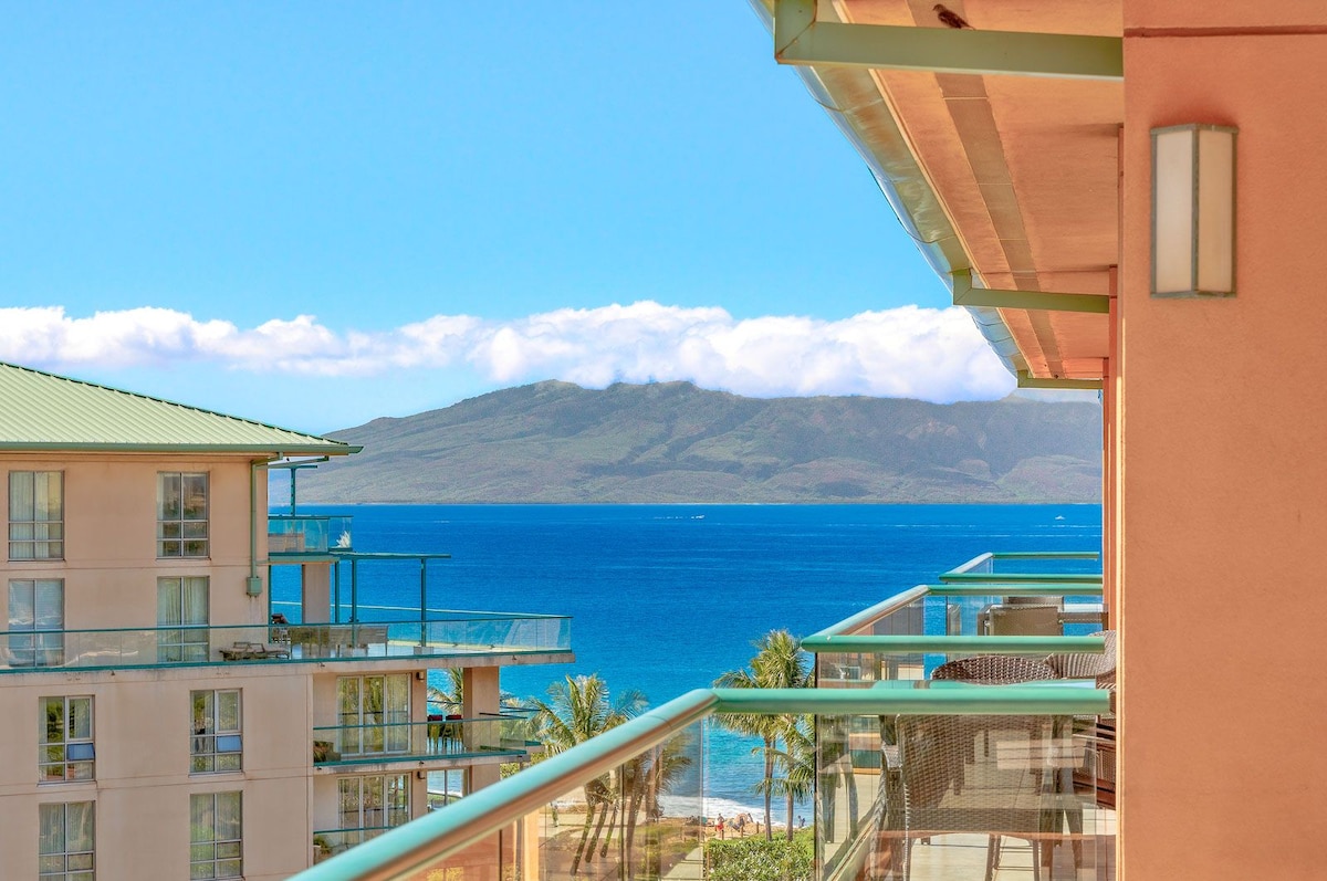 A scenic view shows the blue Pacific Ocean complemented by the West Maui Mountains in the background. The image captures the expansive balcony railing, with sunlight illuminating the area and palm trees swaying gently nearby.