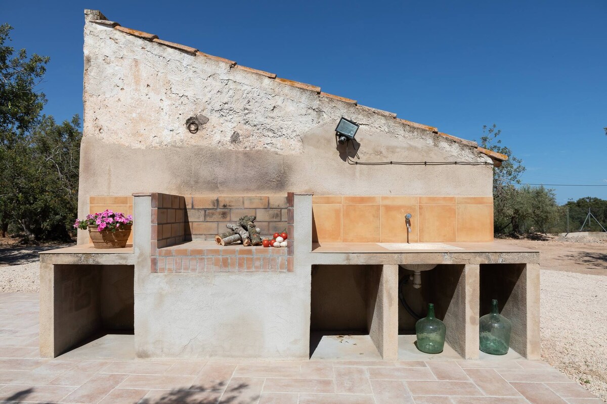 An outdoor barbecue area is featured, constructed with rustic materials. A built-in grill is surrounded by a stone countertop, complemented by a colorful flower pot. Beneath the counter, storage spaces and containers can be seen, against a backdrop of clear blue skies.