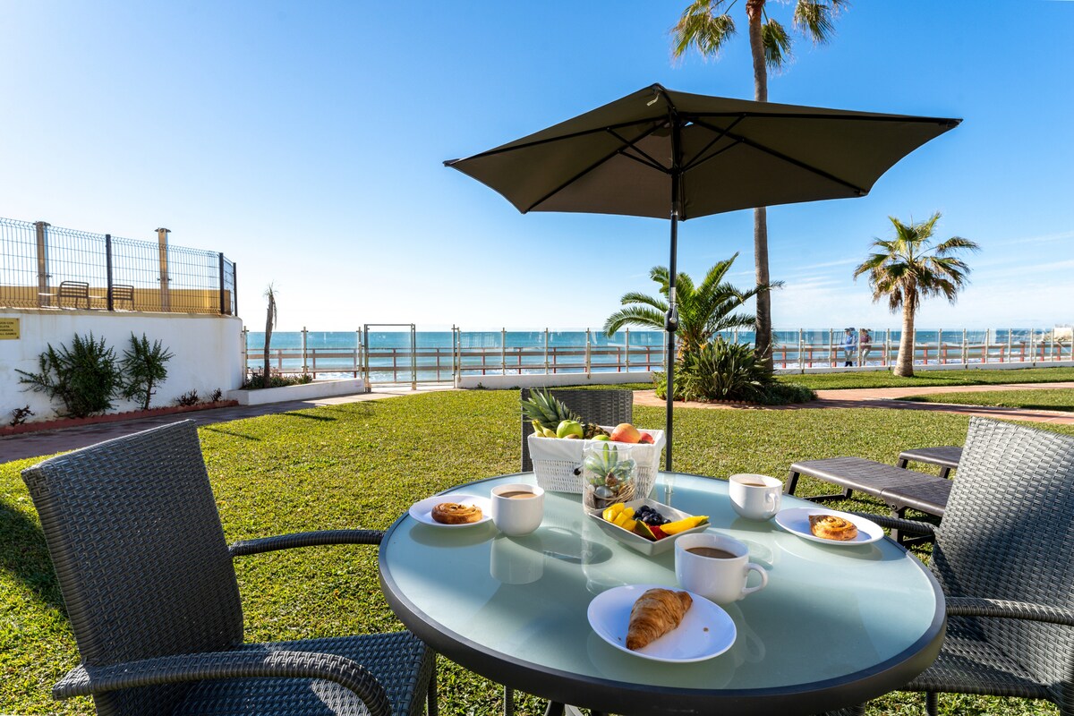 A serene outdoor dining area is presented, featuring a round table with a sun umbrella. Fresh fruit, pastries, and coffee are arranged on the table. In the background, the Mediterranean Sea sparkles under a clear blue sky, bordered by a walking path and palm trees.