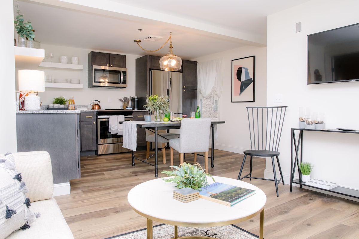 An open-concept area features a modern eat-in kitchen with stainless steel appliances and a dining table for two. A cozy living space includes a light-colored sofa and a flat-screen TV mounted on the wall. Decorative plants and artwork enhance the contemporary design.