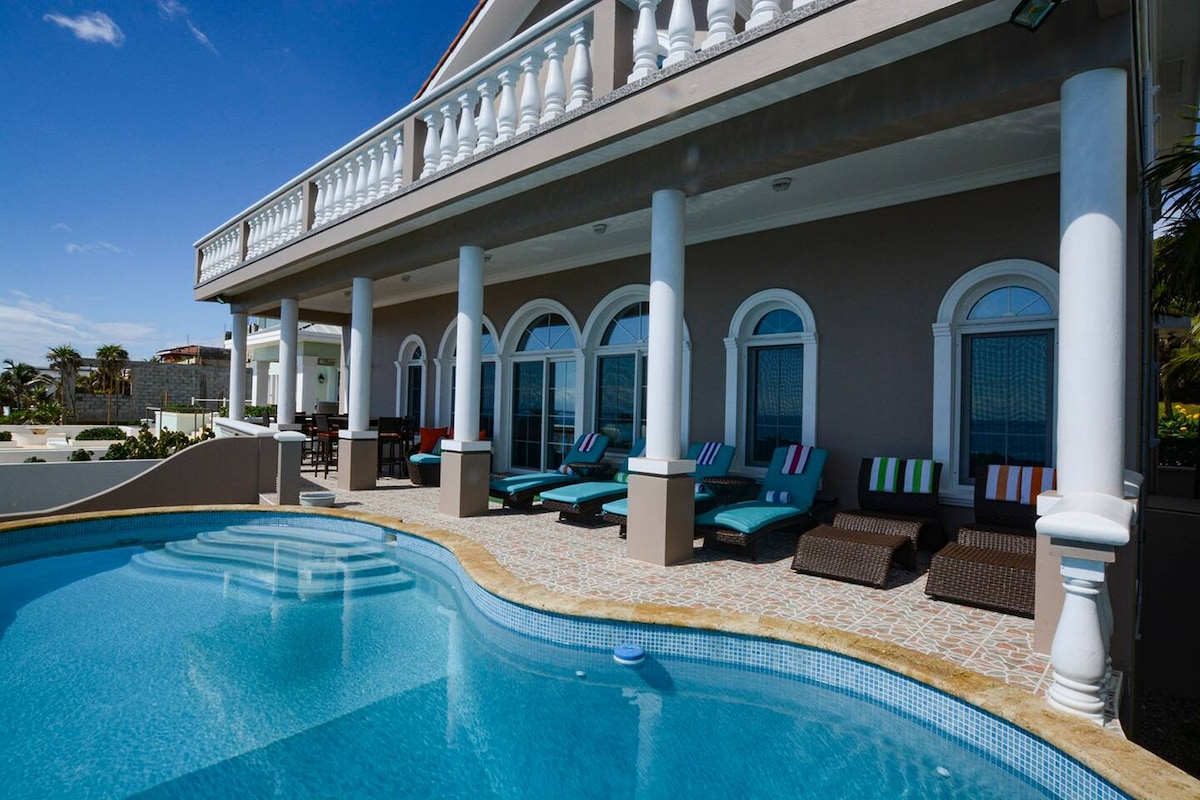 A private pool area showcases a clear, inviting pool bordered by a beautifully tiled deck. Lounge chairs with colorful cushions are arranged neatly beside the pool. The house features wide arches and large windows, enhancing the serene atmosphere against a bright blue sky.