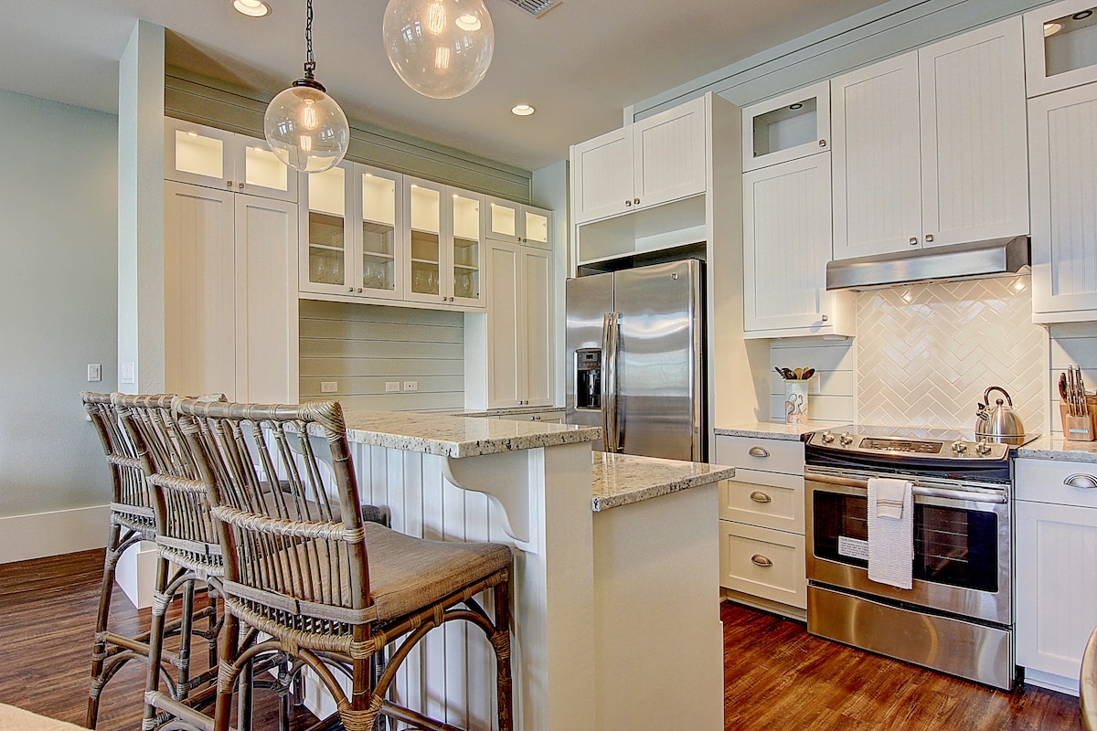The kitchen features light cabinetry with glass inserts, complemented by a marble countertop and stainless steel appliances. A breakfast bar with four wicker stools offers a casual dining option. Subtle tile backsplash enhances the space, and warm wood flooring provides a cozy feel.