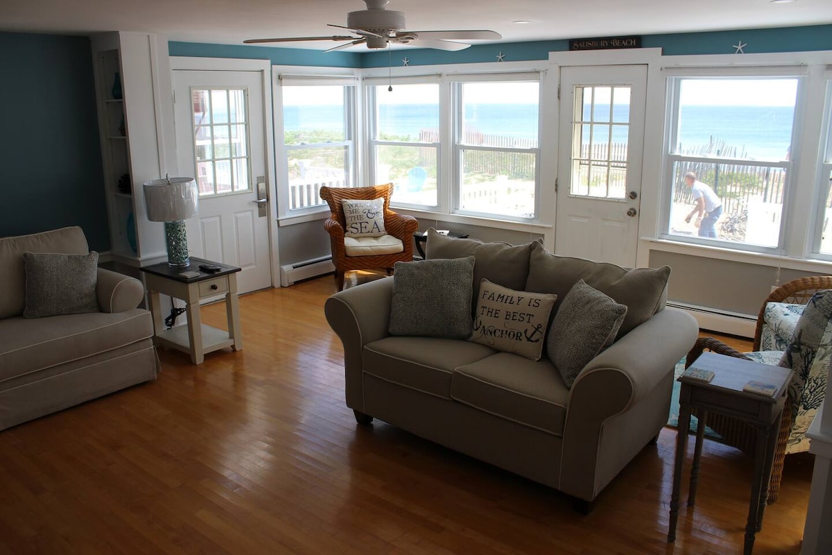 A living room features large windows offering ocean views and allowing natural light to fill the space. Comfortable seating arrangements include a light-colored sofa and armchairs with a coastal theme. The hardwood floor adds warmth to the atmosphere, complemented by a fan overhead.