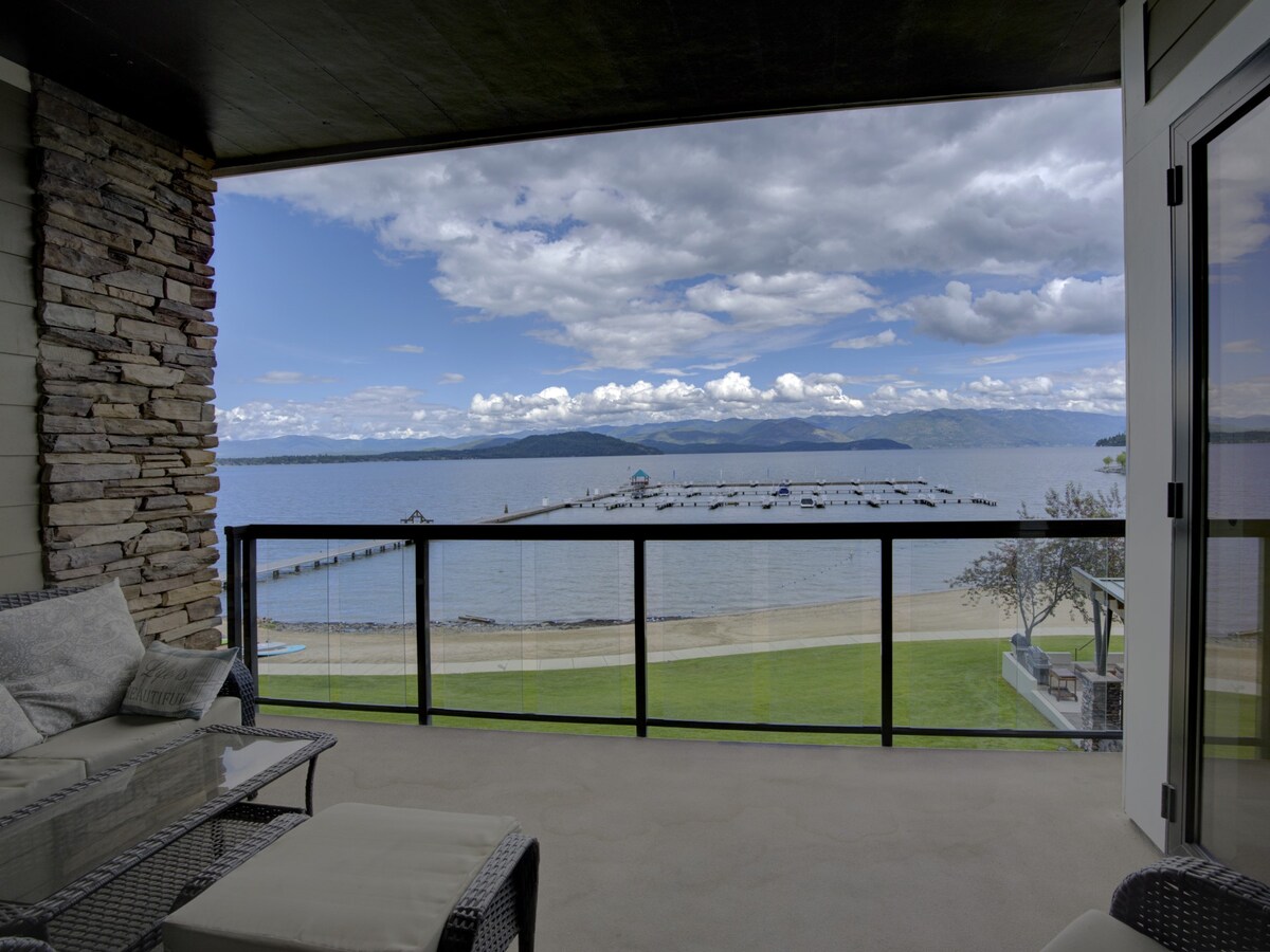 A scenic balcony view features a spacious outdoor seating area overlooking Lake Pend Oreille. The tranquil water reflects the expansive sky dotted with clouds, while a private marina and sandy beach are visible below, framed by lush green grass.