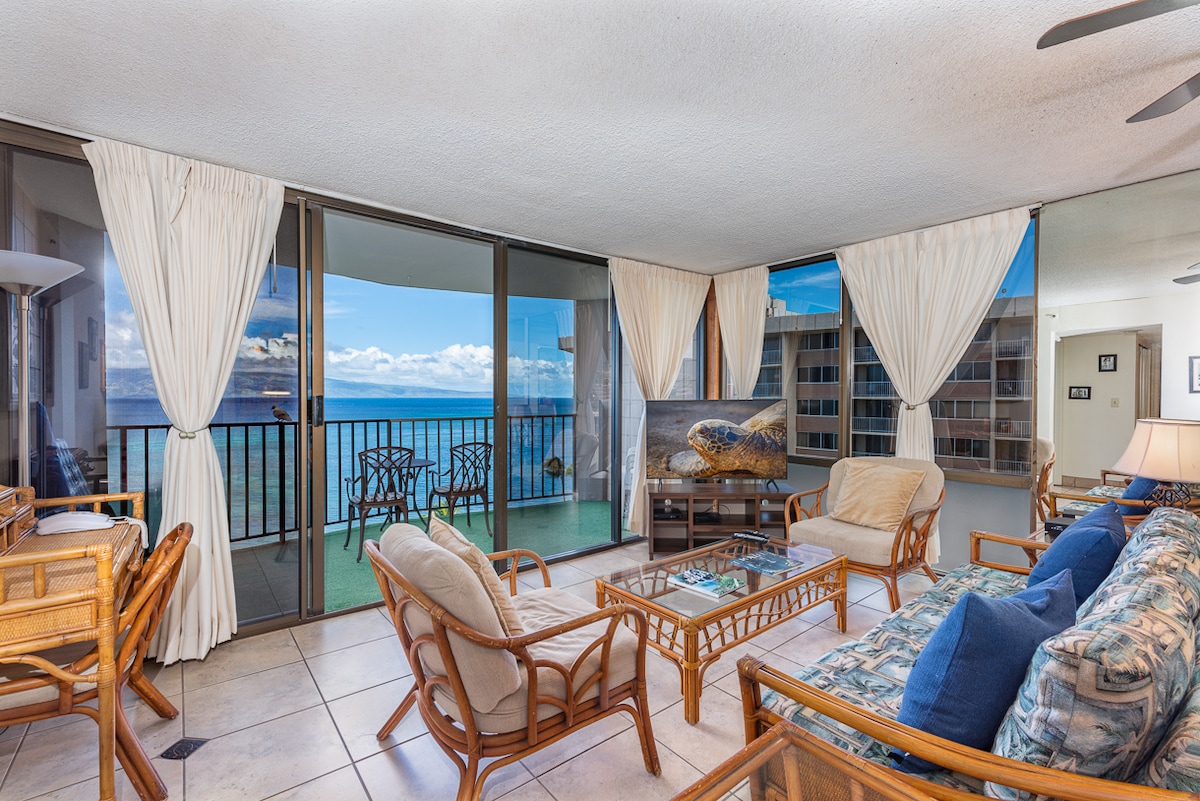 A comfortable living area is presented with rattan furniture, including a spacious sofa and two chairs. Large glass doors open to a balcony with ocean views. Light curtains frame the windows, and a television is positioned on a stand, with decorative items on the coffee table.