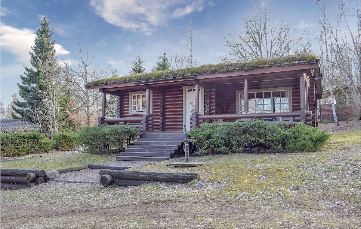 The cozy cottage features a wooden façade with a green moss-covered roof. Steps lead to a welcoming entrance framed by large windows. Surrounding greenery adds to the natural setting, while a gravel pathway and logs are visible in the foreground.