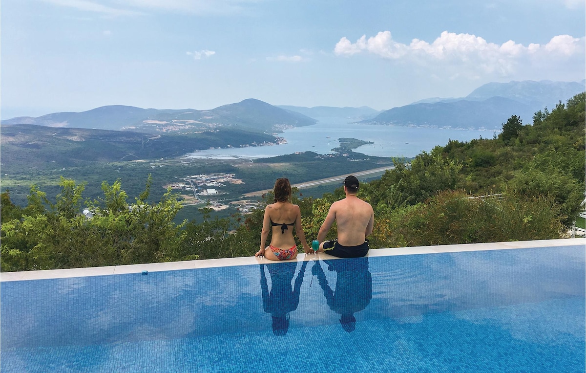A serene scene by the infinity pool, where two individuals sit side by side, enjoying sweeping views of the Bay of Tivat and surrounding mountains. The tranquil water mirrors the clear sky, while lush greenery frames the extraordinary landscape.