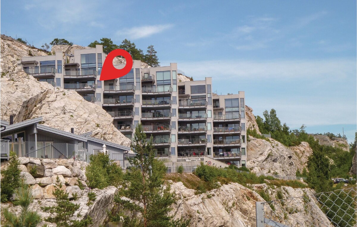 The modern apartment building is situated on a rocky hillside, featuring multiple levels with glass balconies. Green trees are visible in the foreground, and the sky is partly cloudy, creating a serene backdrop for the structure.