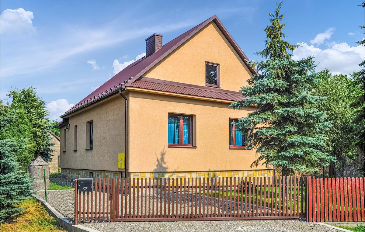 The exterior of the cottage features a light-colored façade with a sloped roof and a welcoming front entrance. Surrounding greenery includes tall trees and shrubs. A wooden fence lines the property, providing a sense of privacy, while a gravel path leads to the front door.