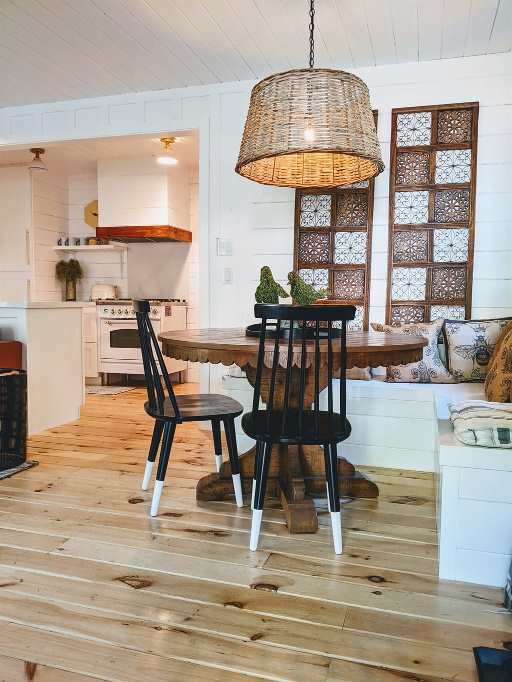 A round wooden dining table is surrounded by four black spindle-back chairs, featuring white-painted legs. Above, a woven pendant light casts a warm glow, complementing the white paneled walls. Decorative panels with intricate patterns add a rustic charm, and cozy cushions are arranged on nearby seating.