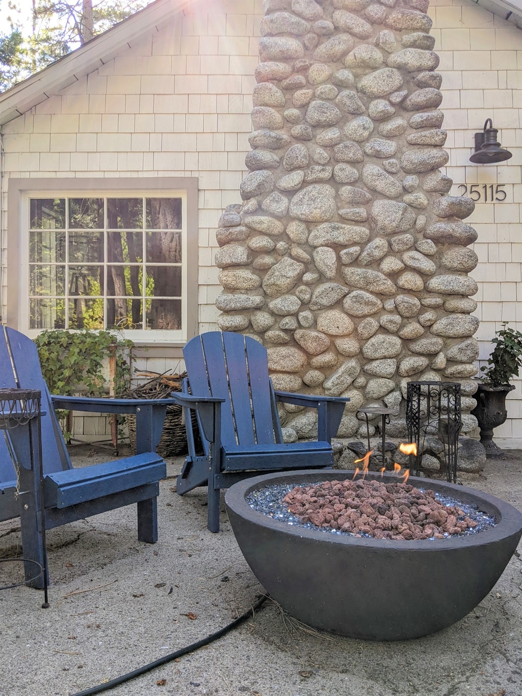 A cozy outdoor seating area is featured with two blue adirondack chairs positioned near a round fire pit, surrounded by pebbles and decorative rocks. A charming stone chimney serves as a backdrop, with a window showcasing the inviting interior of the cottage.
