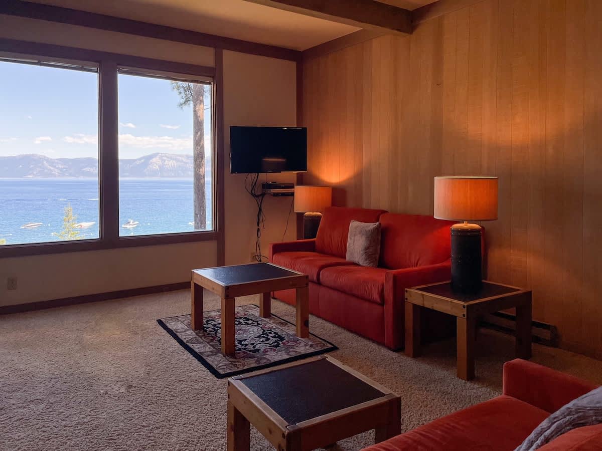 A cozy living area features two red sofas, accompanied by wooden coffee tables. Large windows offer expansive views of the lake and surrounding mountains, while a television and soft lighting create a comfortable atmosphere. The carpeted floor adds to the warmth of the space.