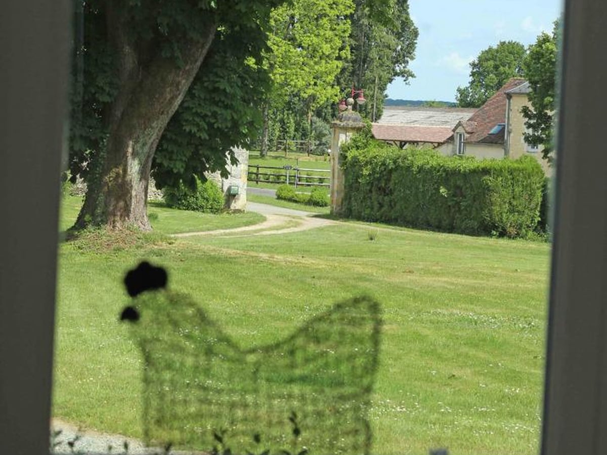 A view of the serene landscape is seen through a window. Lush green grass stretches across the foreground, framed by trees. A winding path leads toward an entrance gate, while a few farm buildings are visible in the background, surrounded by greenery.