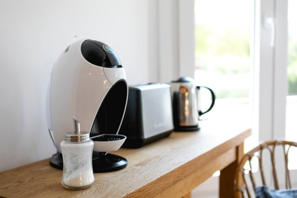 A sleek coffee maker, kettle, and toaster are arranged on a wooden countertop. Sunlight filters through a nearby window, highlighting the clean lines of the appliances and the simplicity of the design. A small sugar dispenser rests beside the coffee maker.
