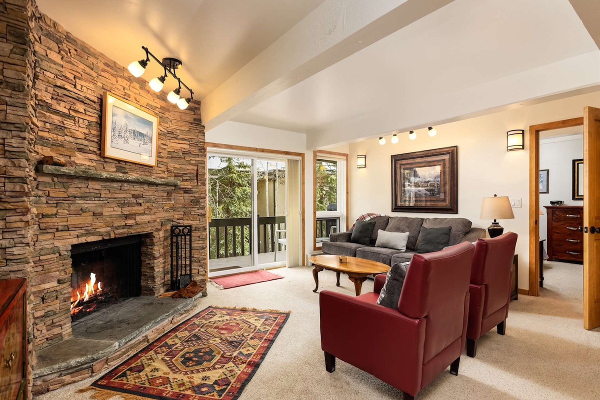 A living room with a stone fireplace creates a cozy focal point. Comfortable seating is arranged around a wooden coffee table, while light floods in through large windows. A patterned rug lies beneath, enhancing the warm ambiance of the space.