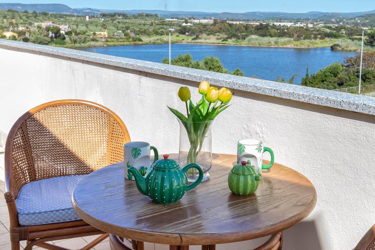 A round wooden table is set with a green tea kettle, two mugs, and a vase of yellow tulips. A wicker chair complements the arrangement. The background showcases a panoramic view of the lagoon and lush landscape beyond.