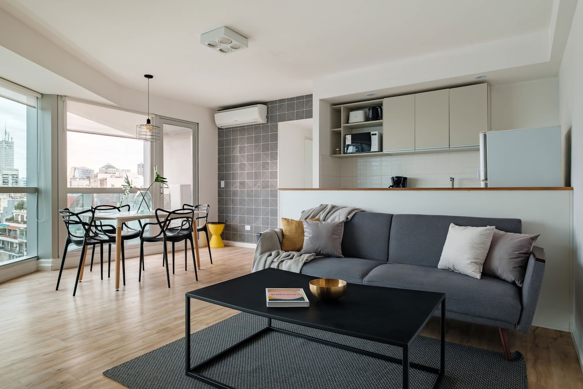 A bright living area features a comfortable sofa accompanied by decorative cushions. A sleek coffee table sits atop a textured rug, and large windows display a city view. The dining space includes a round table with four chairs, illuminated by natural light.