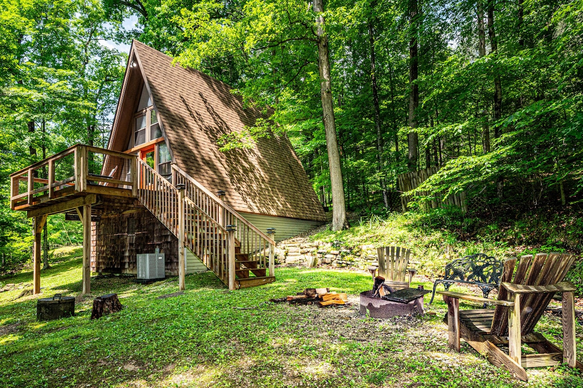 The A-frame chalet is nestled among tall trees, featuring a wooden deck that provides access to the entrance. A fire pit surrounded by several seating options is present in the grassy yard, inviting relaxation in the serene woods.