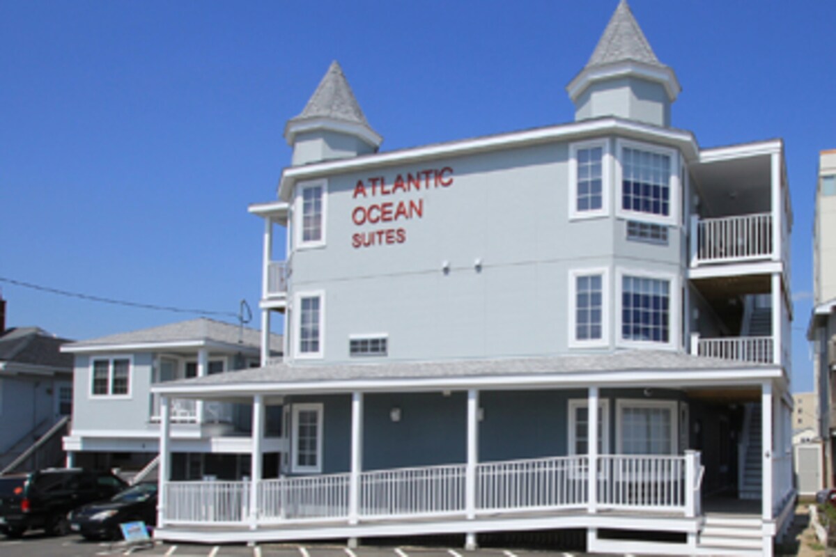 The exterior of Atlantic Ocean Suites is presented, featuring a two-story building with light blue siding and peaked roofs. Large windows provide ample natural light, and a wide porch with white railings invites outdoor relaxation. Parking spaces are visible in front of the property.