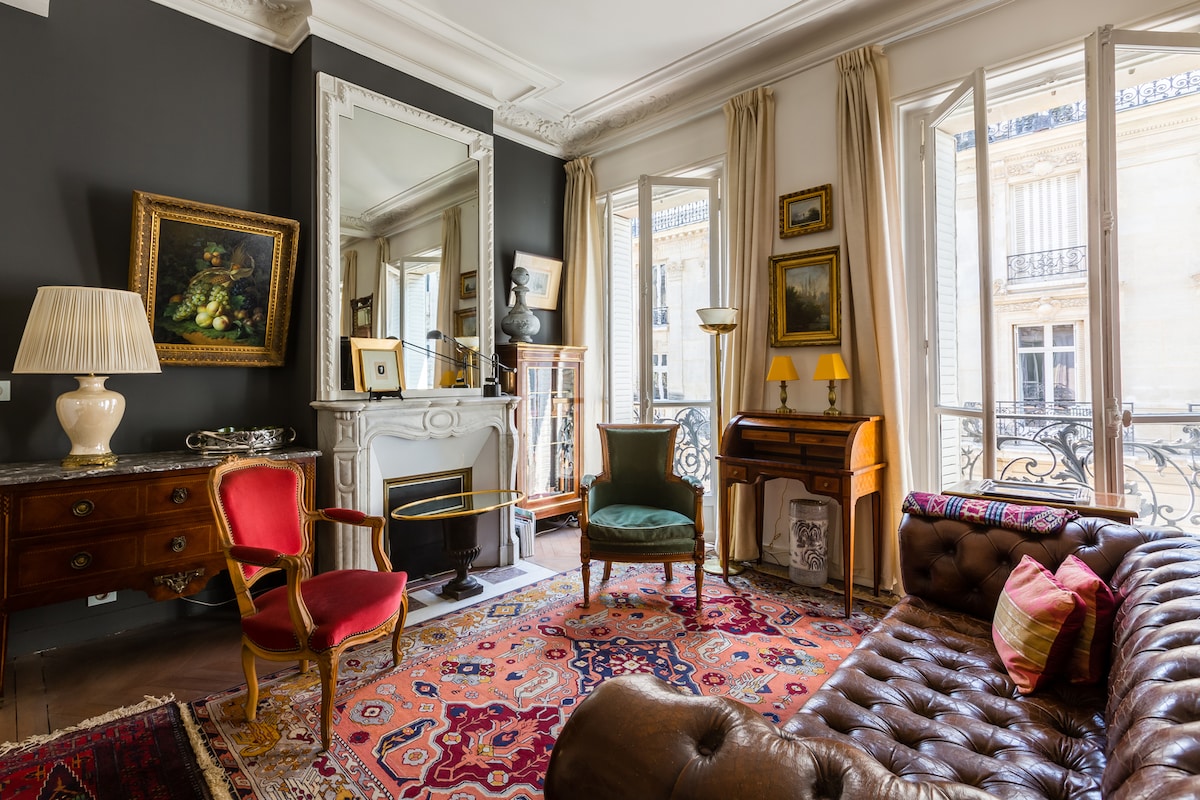 The living area features classical elements such as corniced ceilings and wood beams. A plush leather sofa is complemented by an assortment of patterned rugs. Decorative antique chairs, an ornate mirror, and a wooden console table add character, with light streaming in through large French doors.