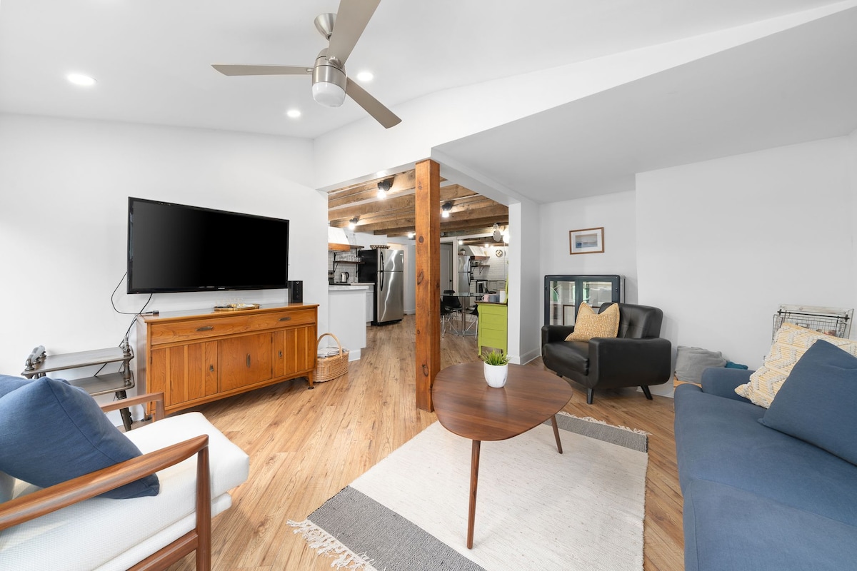 The living area features wooden flooring and exposed beams, creating a modern yet warm ambiance. A large flat-screen TV is mounted on one wall, while a wooden sideboard and a small coffee table are positioned in the center. Comfortable seating options include a dark leather chair and a blue fabric sofa, complemented by decorative pillows.