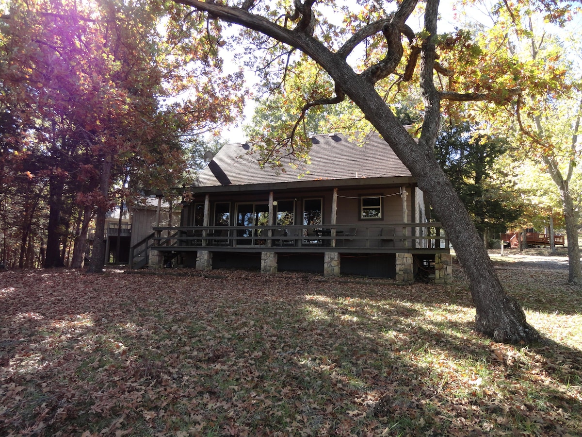 A lakefront cabin is set amidst a tranquil wooded area, surrounded by colorful autumn foliage. The structure showcases a covered front porch supported by stone pillars. Large windows provide views into the interior, and a level lawn leads to the edge of the serene environment.