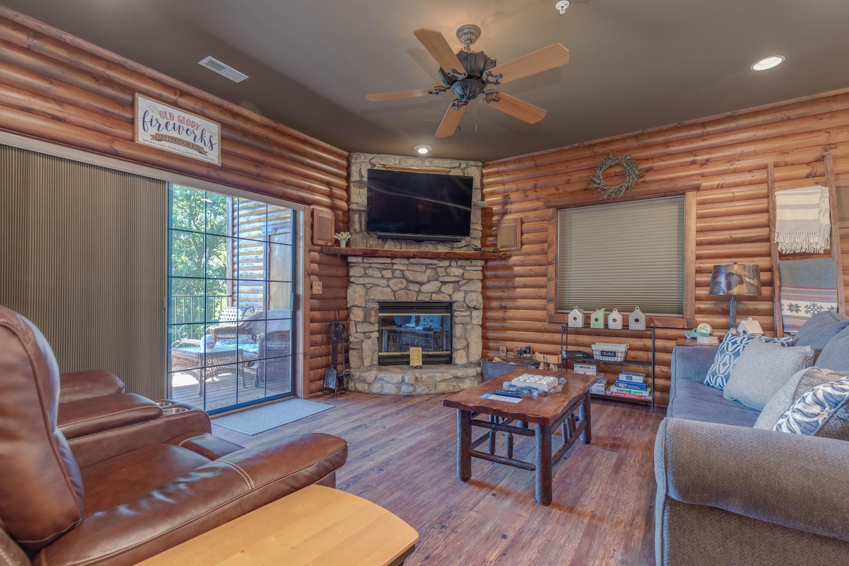 A log cabin living area features a stone fireplace with a mounted television above. Dual recliners and a plush sofa offer seating options. Natural light filters through sliding glass doors that access a deck, enhancing the space's inviting ambiance.