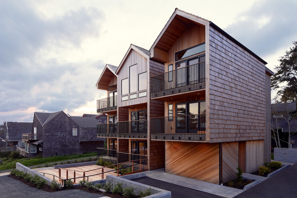The exterior view of a multi-level building showcases a contemporary design with wooden siding and expansive balconies. The structure is flanked by landscaped areas, and a shared deck area is visible in the foreground, suggesting space for relaxation and gatherings.