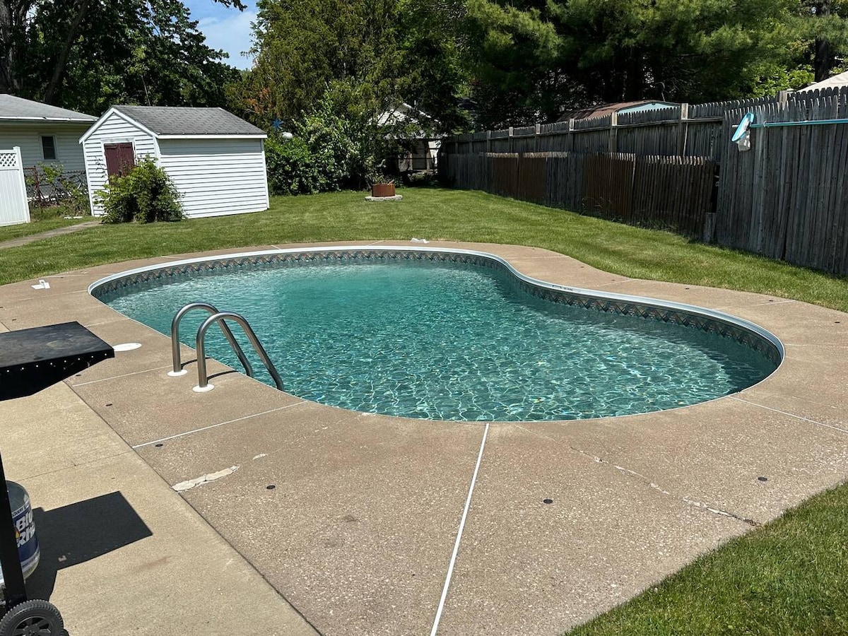 A private in-ground pool is surrounded by a spacious concrete deck, with clear water reflecting sunlight. Lush green grass borders the pool area, and a privacy fence encloses the backyard. A neighboring shed is visible in the background, adding to the outdoor space.
