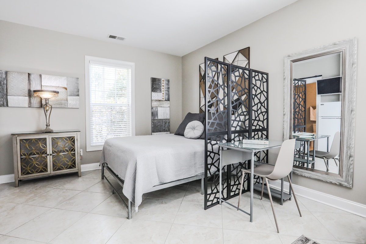 A spacious studio bedroom features a queen size bed positioned against a modern accent wall. A desk and chair are situated beside a decorative room divider. Natural light filters through a window adorned with blinds, while a mirrored dresser adds depth to the serene space.