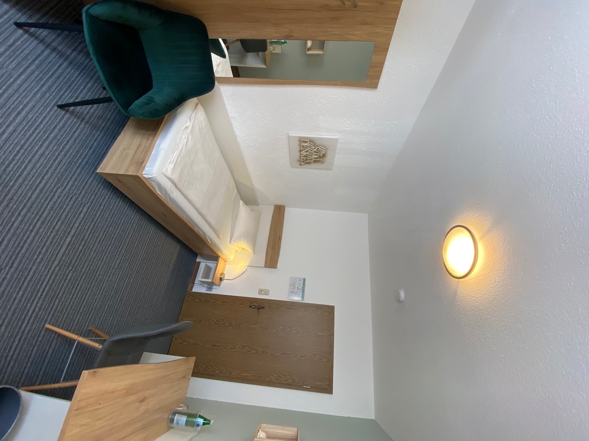 A single room is seen with a spacious bed placed against the wall. A simple wooden desk and an inviting green chair complement the room's decor. A mirror reflects the light from the overhead fixture, enhancing the bright, airy ambiance.