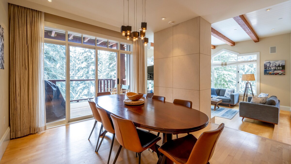 A spacious dining area is highlighted by a large round table surrounded by modern chairs. Large glass doors provide access to an outdoor balcony, offering views of the surrounding trees. Natural light filters in through the windows, enhancing the inviting atmosphere.