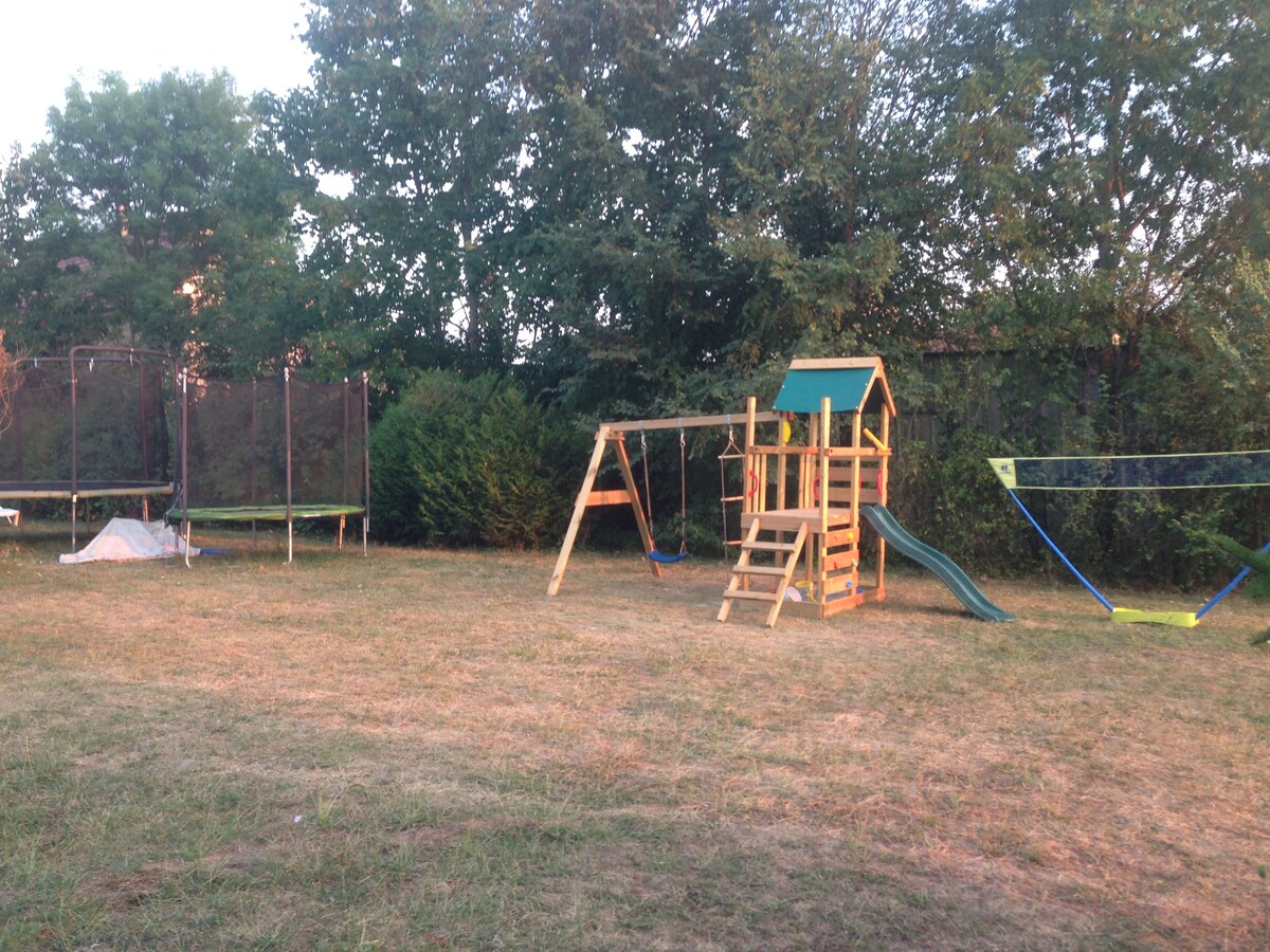 An outdoor play area is depicted featuring a wooden playground structure with a slide and swings. A trampoline is visible nearby, surrounded by grassy patches and trees, providing shaded spots. The setting is spacious, allowing for recreational activities in a natural environment.