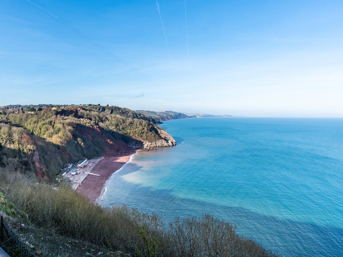 A panoramic view of a calm beach, featuring soft, sandy shores that meet tranquil turquoise waters. Lush greenery lines the coastline, with distant hills providing a scenic backdrop under a clear blue sky. The overall scene conveys a sense of serenity and natural beauty.