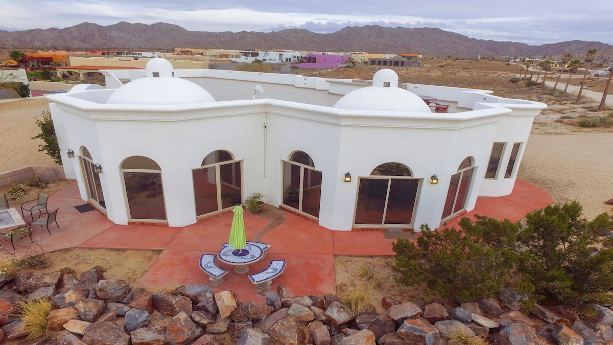 A spacious white house with distinctive dome roofs is surrounded by rugged desert landscaping. The exterior features large arched windows and a patio area, which includes a decorative stone compass design and a vibrant green umbrella for shade against the backdrop of mountains.