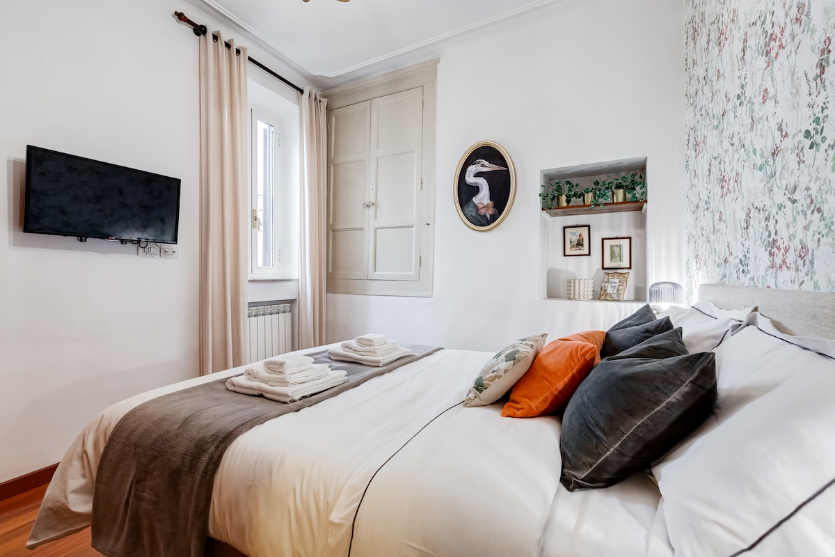 The bedroom features a spacious bed adorned with decorative pillows and fresh linens. A flat-screen TV is mounted on the wall opposite. Natural light enters through a window, and wall shelves display decorative items, enhancing the serene atmosphere.