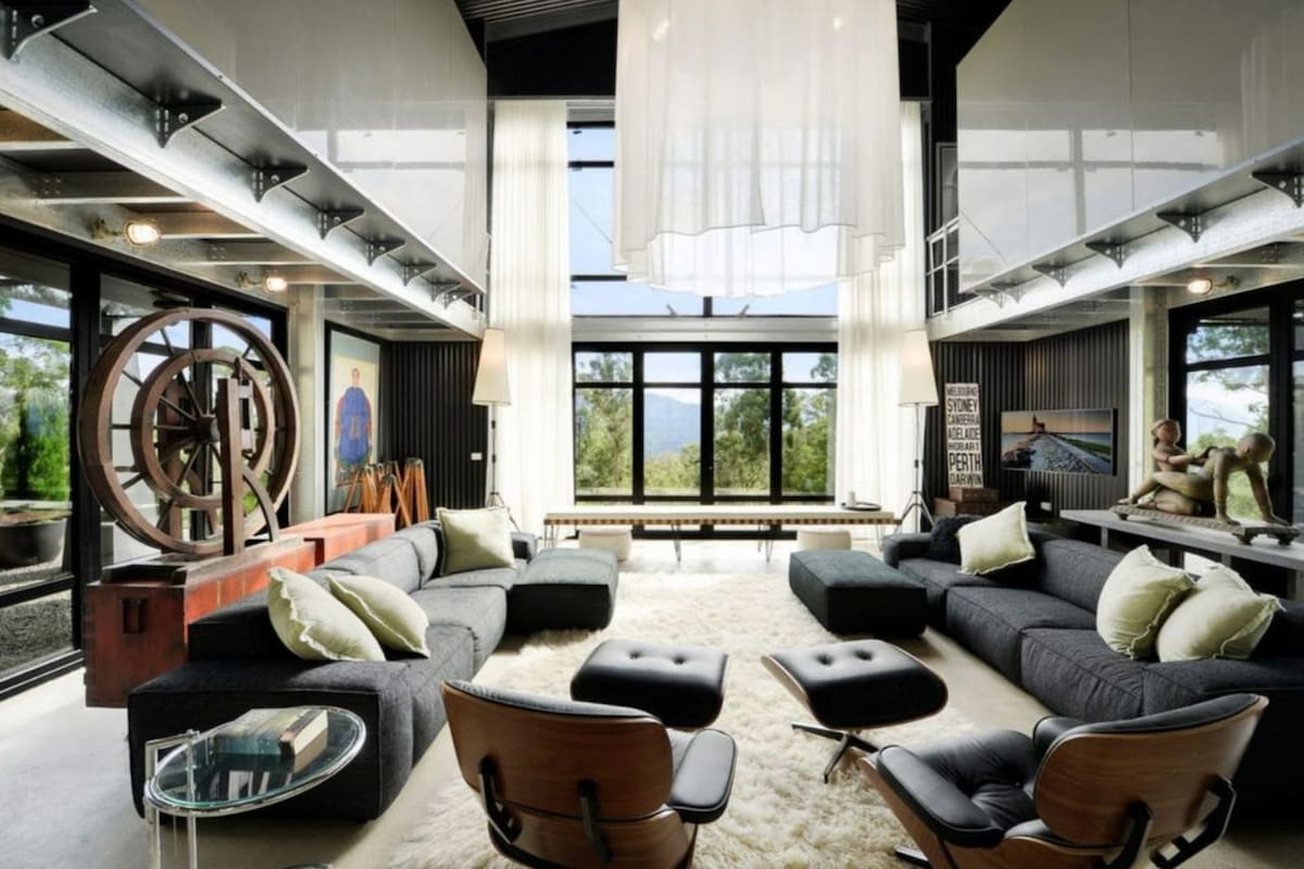 The spacious open-plan living area features large glass windows that invite abundant natural light and offer views of the surrounding landscape. Industrial elements are highlighted by artwork and distinctive furnishings including a low-profile sectional sofa, accent chairs, and a central glass table atop a soft area rug.