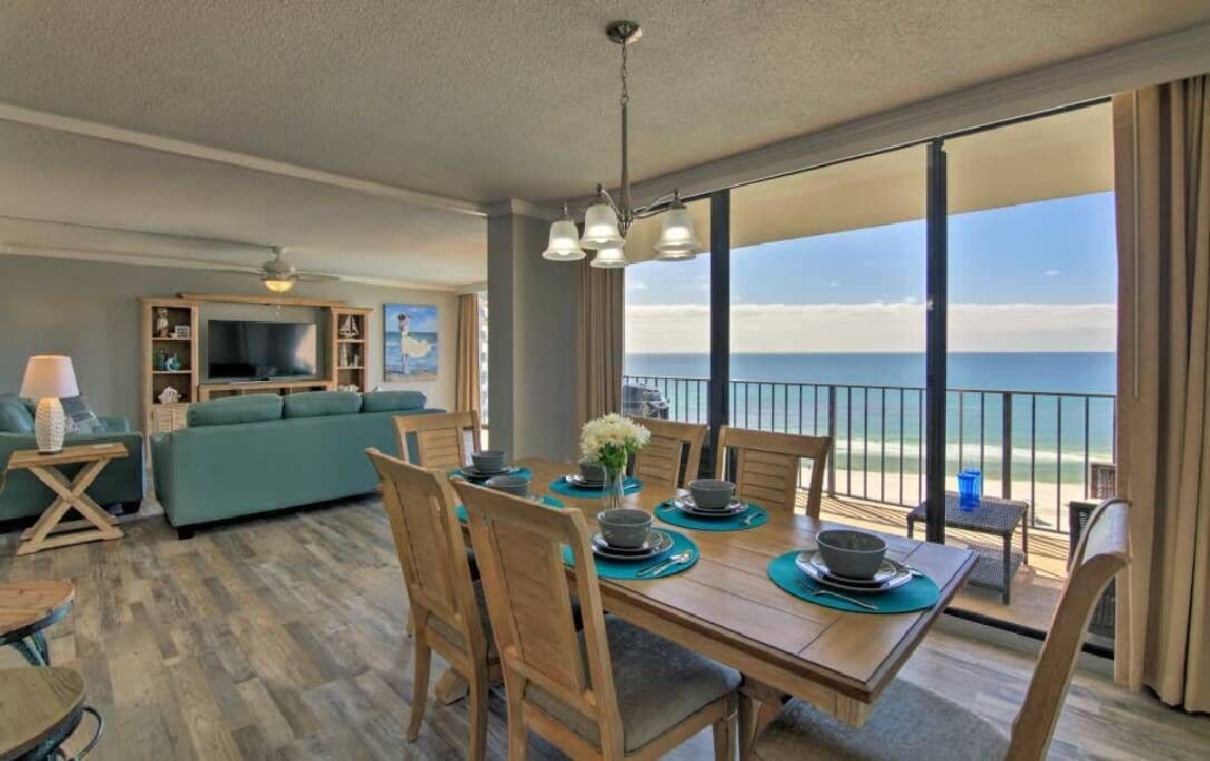 The dining area features a wooden table set for six with coordinating dinnerware. Natural light floods the space through large sliding doors, revealing ocean views just beyond the private balcony. A comfortable seating arrangement is positioned in the background, offering a relaxed atmosphere.
