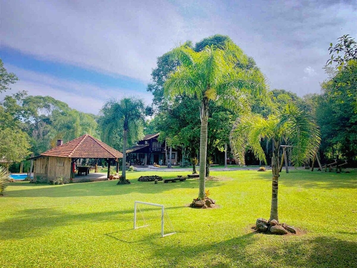 A spacious green lawn is framed by palm trees, offering a serene environment. In the background, a wooden gazebo with a thatched roof provides shade. The main house features a rustic façade, surrounded by a natural landscape, creating an inviting outdoor area.