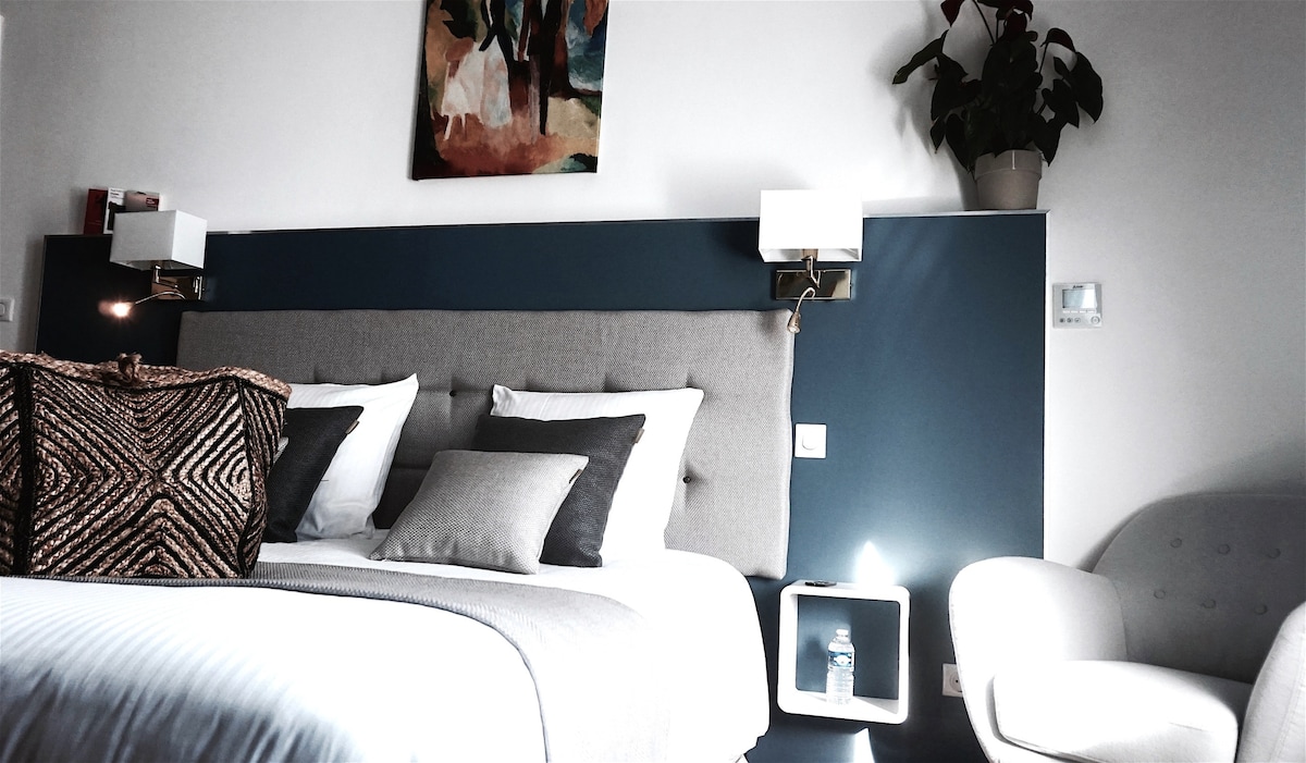 A softly colored bedroom features a spacious bed with decorative pillows against a dark accent wall. A piece of artwork decorates the wall above the bed. A reading lamp is mounted beside the bed, and a cozy armchair is positioned nearby.