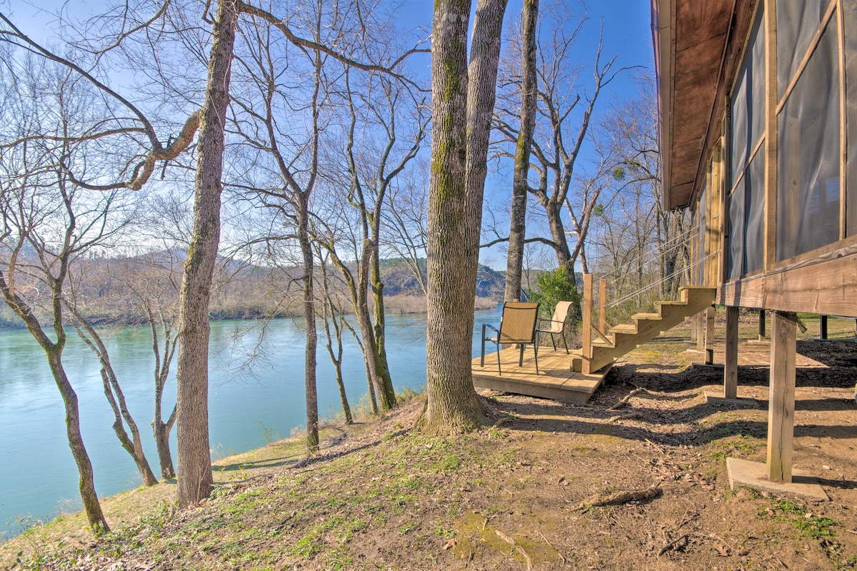 A rustic cabin is set against a riverfront backdrop, surrounded by leafless trees. A staircase leads from the deck down to the riverbank, where seating is positioned to enjoy the tranquil water views. The scene conveys a serene outdoor setting ideal for relaxation.