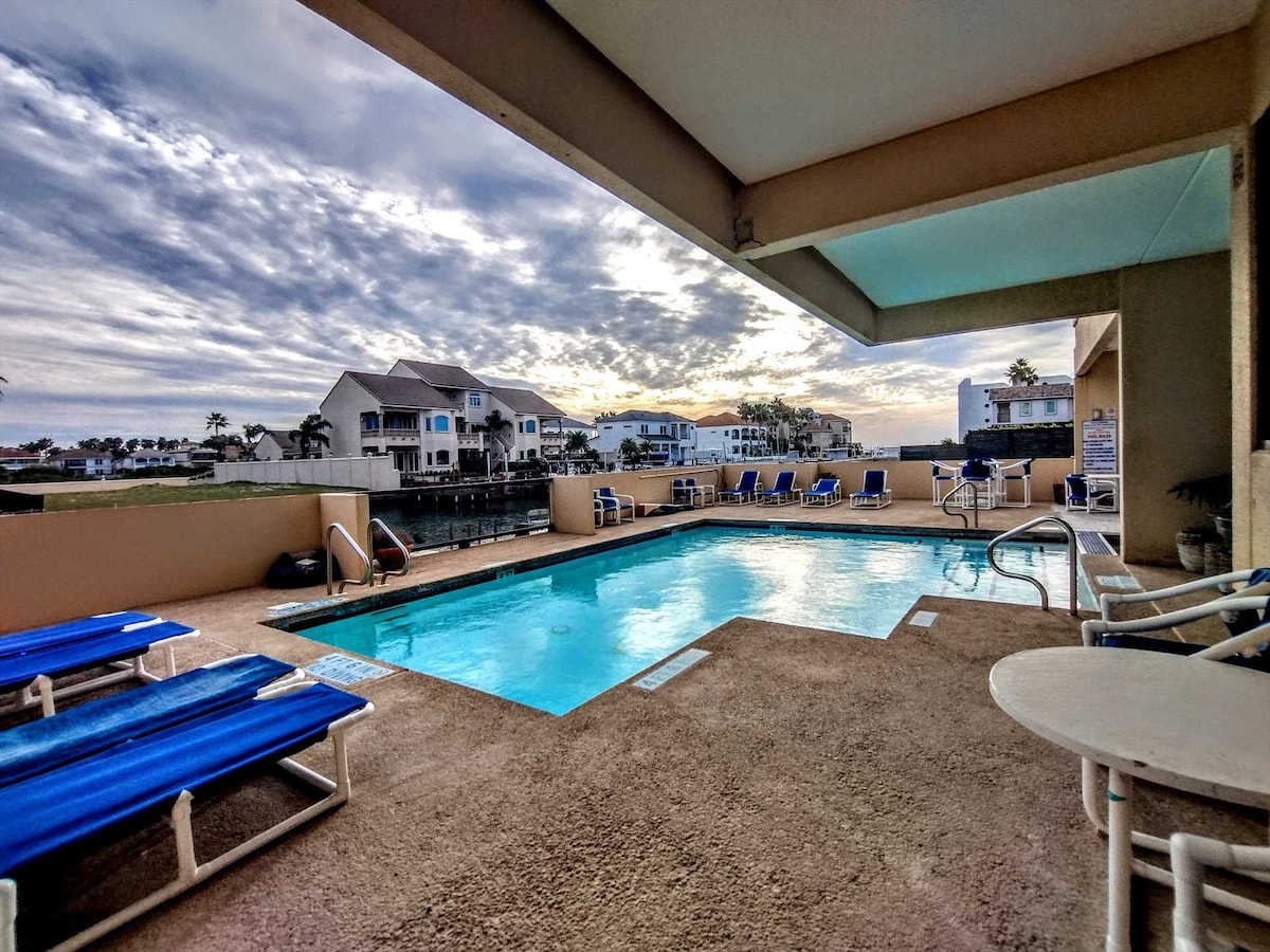 An outdoor pool area features a sparkling blue pool surrounded by lounge chairs. The scene is accented by an evening sky, with buildings and palm trees visible in the background. A small table can be seen near the pool, creating a space for relaxation.
