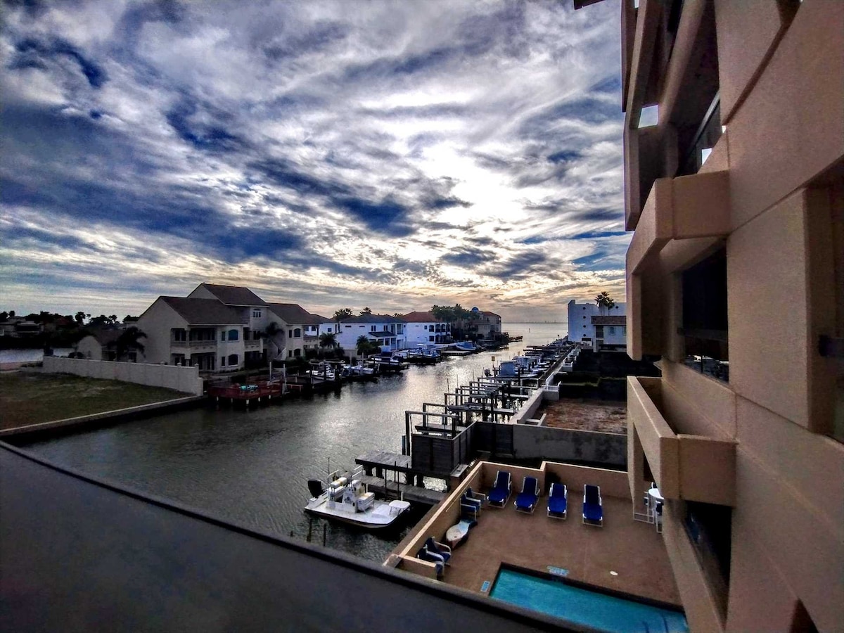 The view from the balcony reveals a peaceful marina lined with boats, reflecting the soft light of the clouds above. A pool area with lounge chairs is visible in the foreground, offering a pleasant spot to relax.